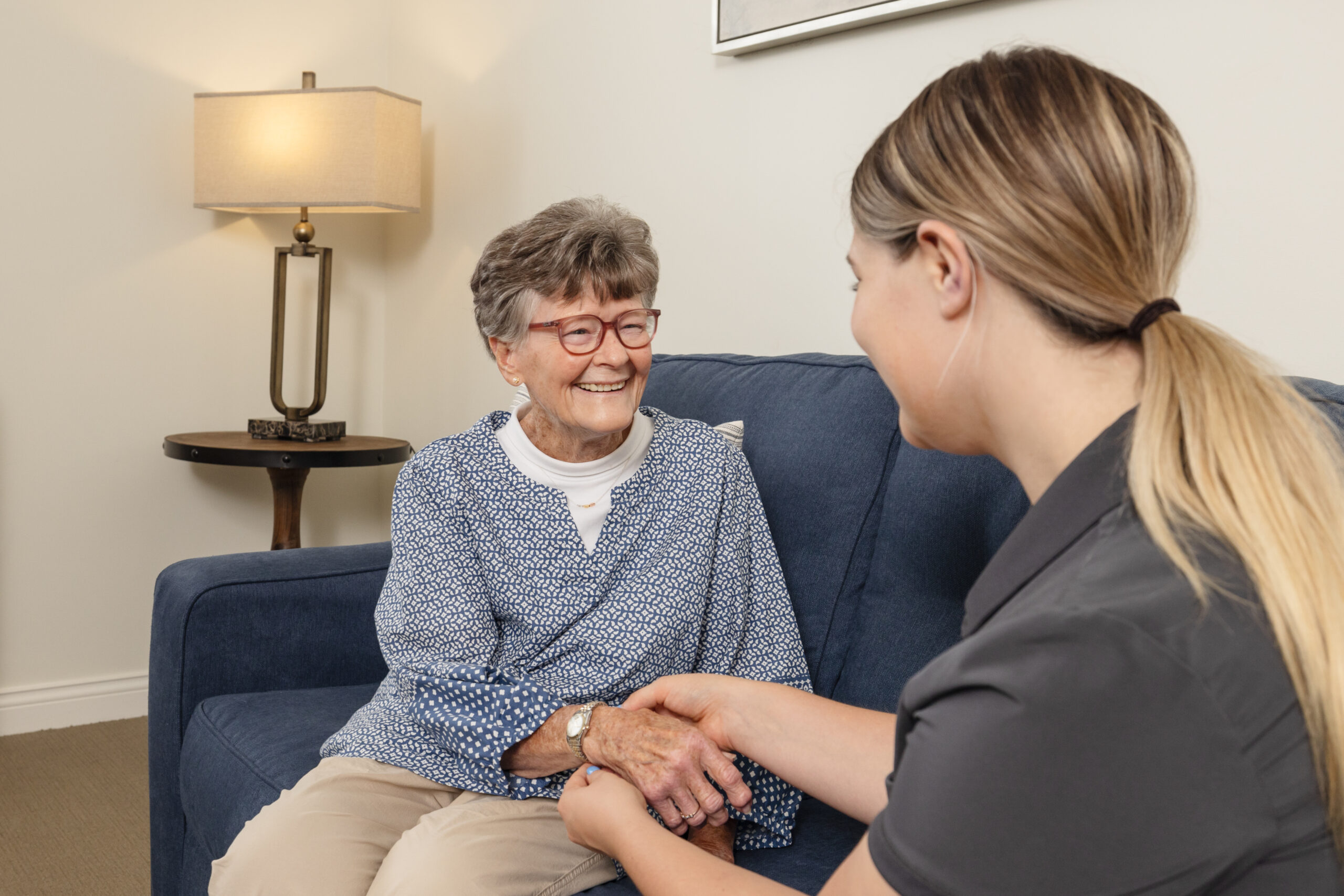 Caring staff member holding hands and chatting with a smiling elderly resident on a blue couch in a welcoming senior living community setting.