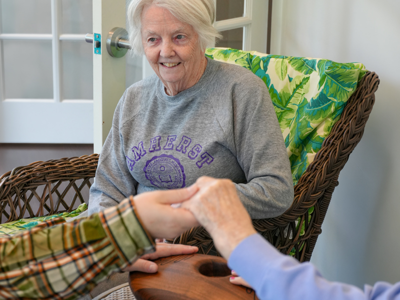 Senior woman in a gray sweatshirt smiles while resting her hands on the Crdl, a TouchPoint Sensory device, with a caregiver holding her hands