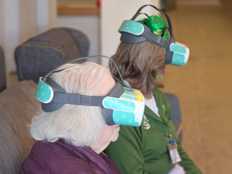 Two senior women wearing virtual reality headsets sit side by side, one in a festive green hat, enjoying an immersive TouchPoint Sensory experience