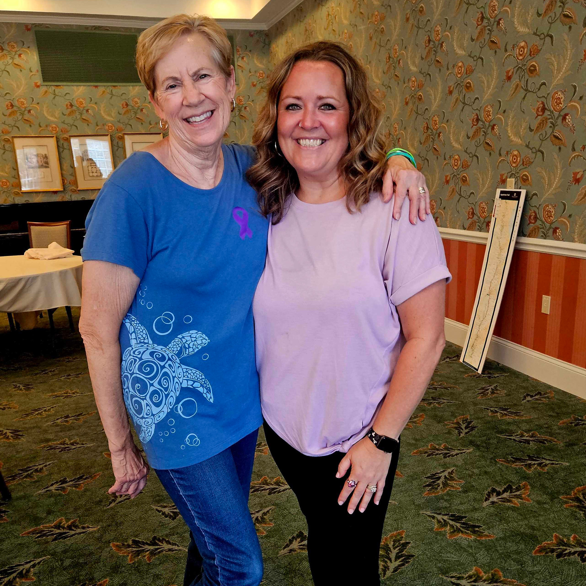Two women wearing purple awareness ribbons smile together inside Berkeley Square during the Memory Care Walk event