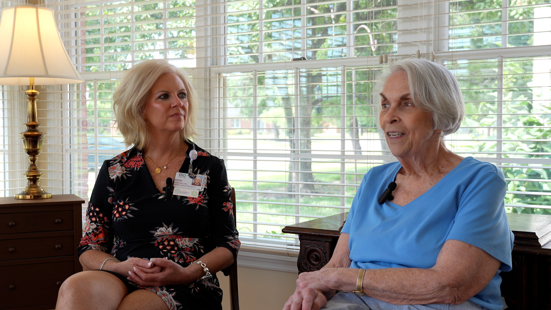Staff member and resident Mary Lee Johns sit together wearing microphones for an interview, chatting in a sunlit room with large windows