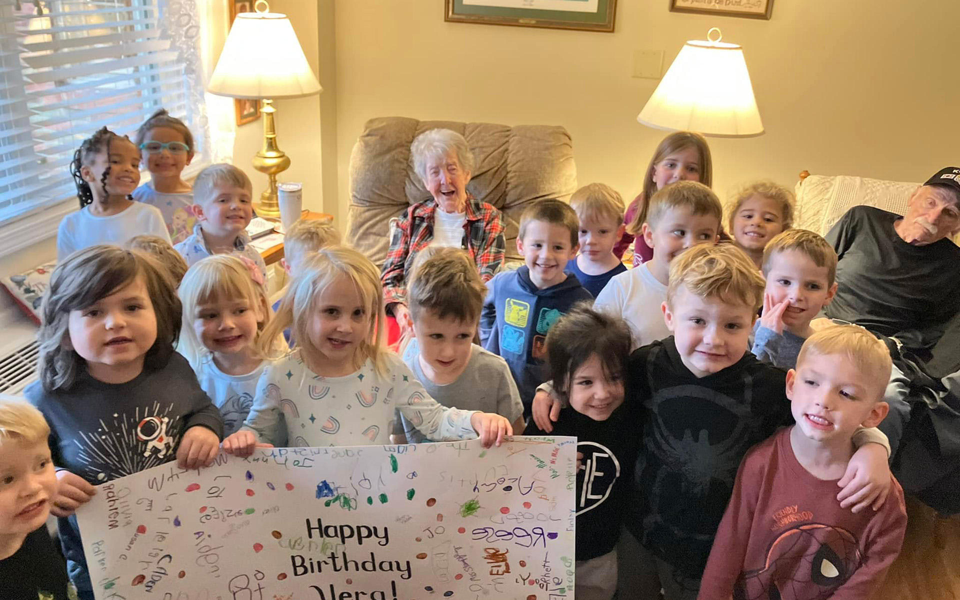 A group of young children surrounds a smiling older resident in her apartment, holding a handmade Happy Birthday sign covered in their signatures.